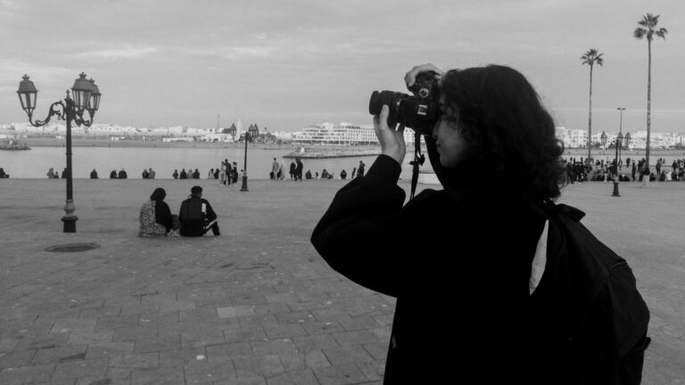 A photographer captures a Moroccan cityscape with people and palm trees beside the waterfront.