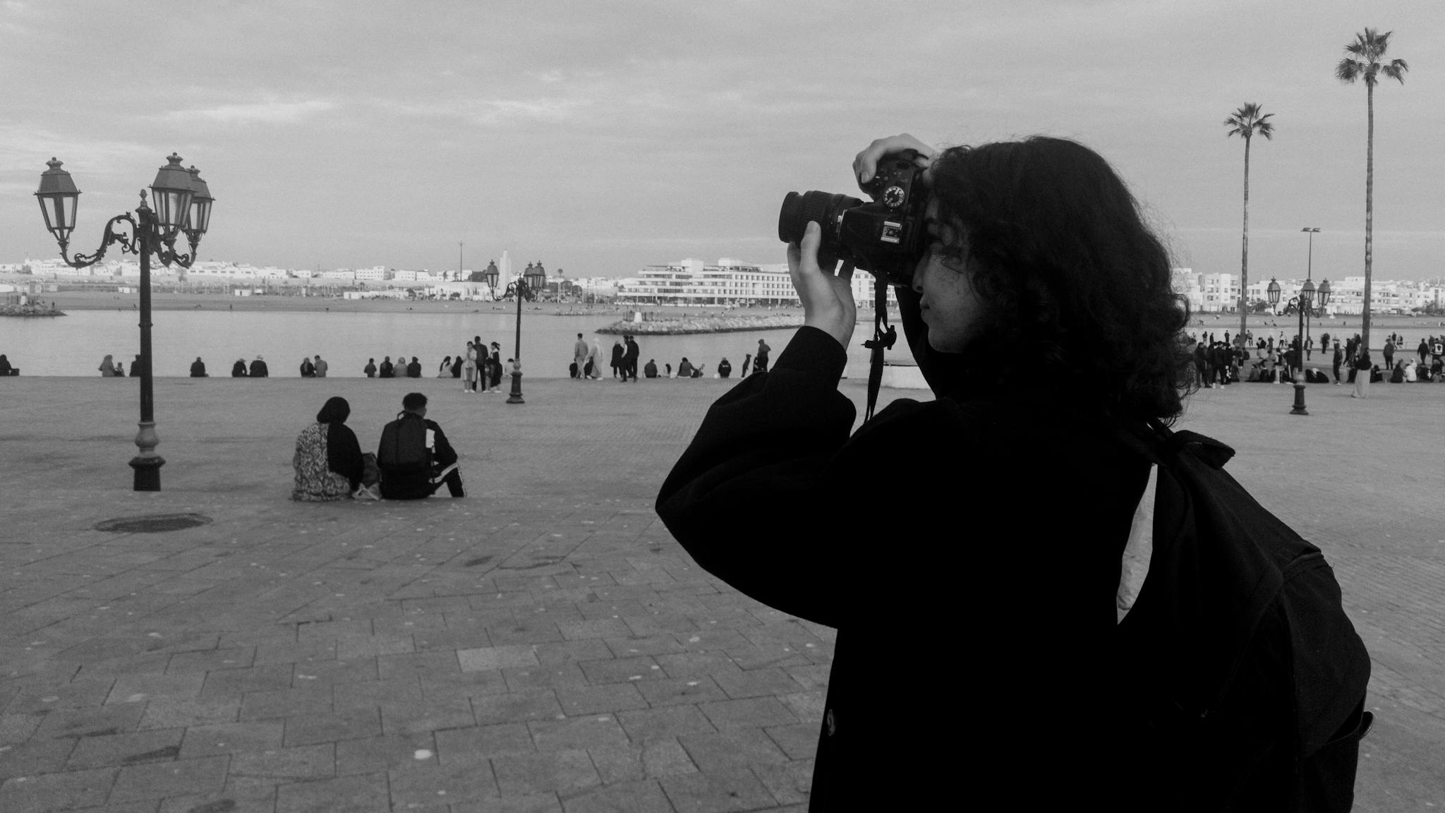 A photographer captures a Moroccan cityscape with people and palm trees beside the waterfront.