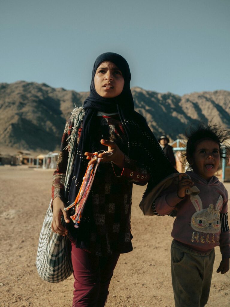 Two children walking together in Dahab, Egypt, with mountainous desert backdrop under clear skies.